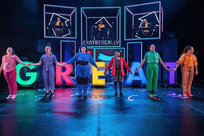 Group of women wearing brightly coloured clothing holding hands on a stage at the Edinburgh Festival Fringe